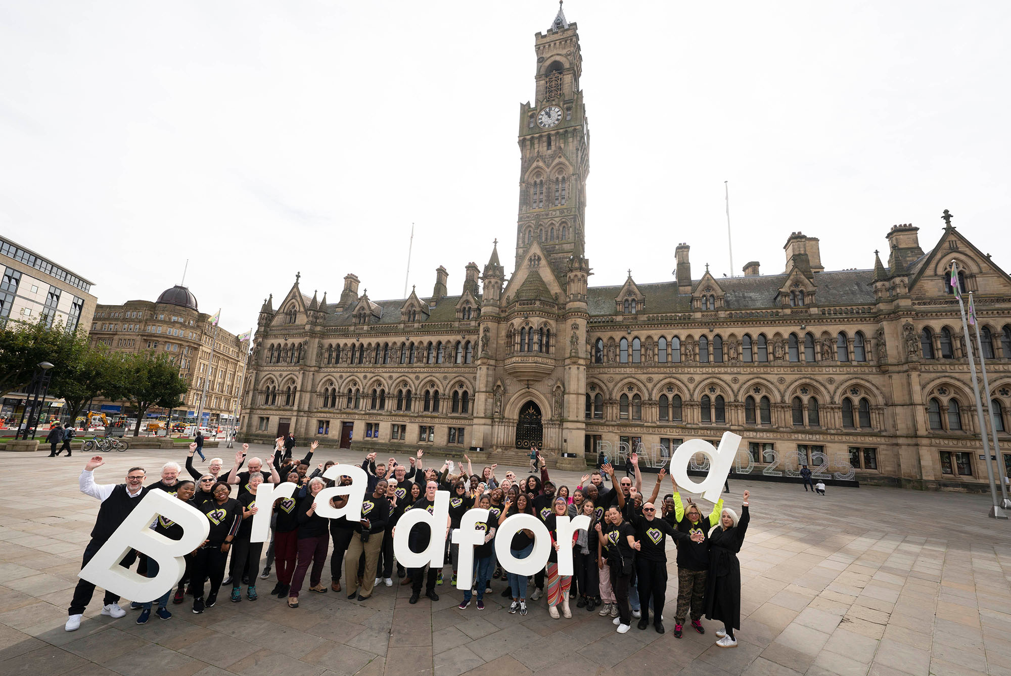 A group of people holding large letters spelling the word BRADFORD, standing in front of a large, old stone building