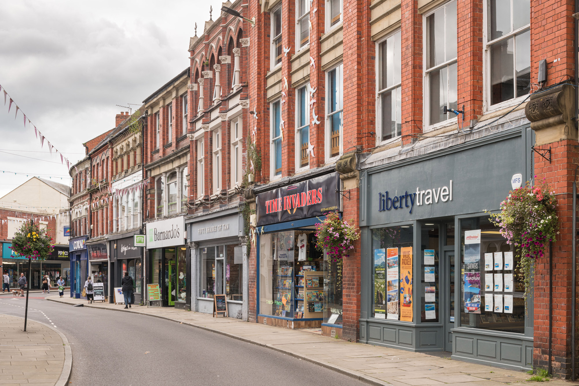 A colour photo showing a high street winding into the distance. Some pedestrians are making their way along pavements. 