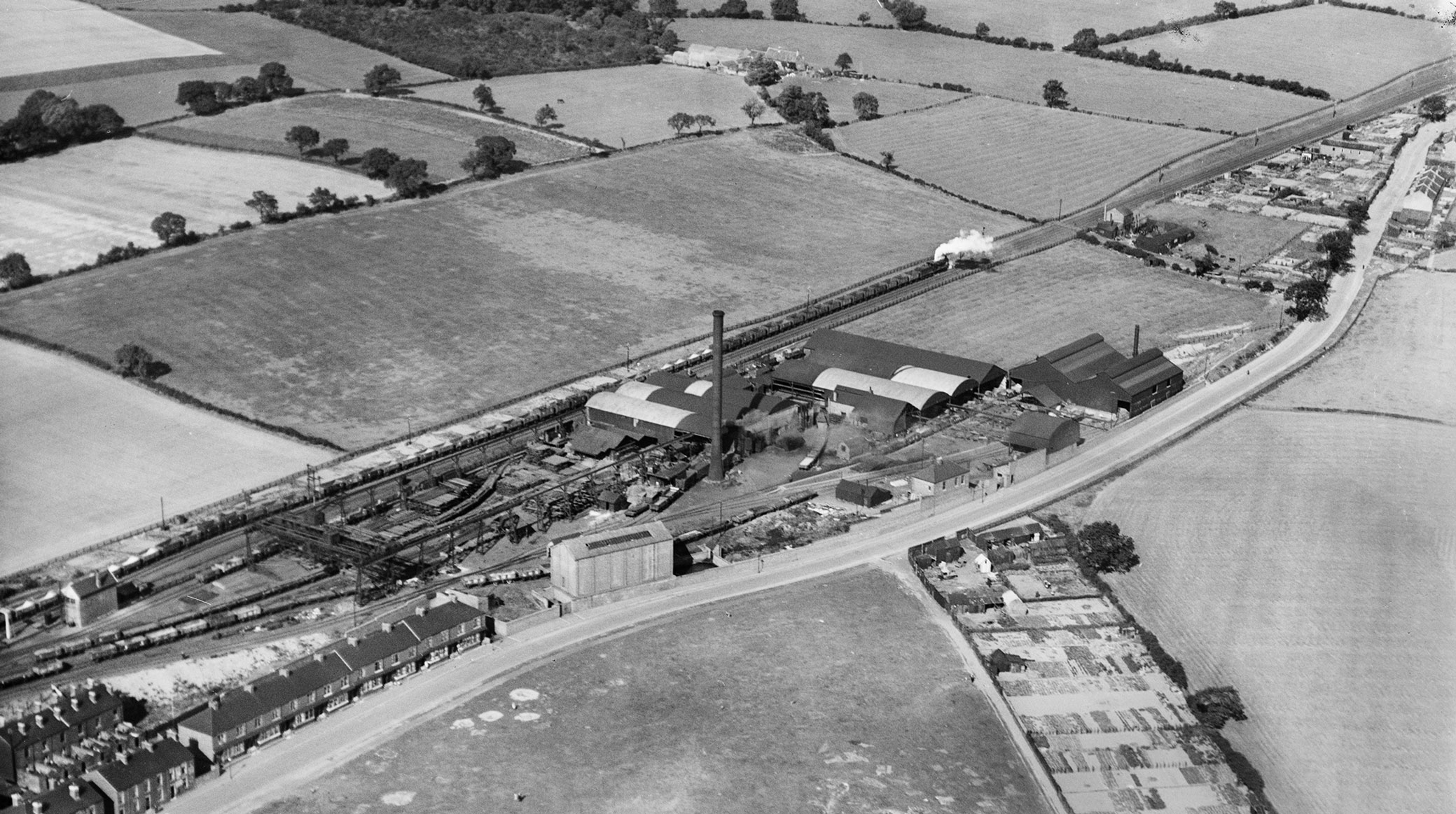 A black and white aerial  photograph showing a steam freight train moving past a small industrial complex surrounded by a patchwork of fields.
