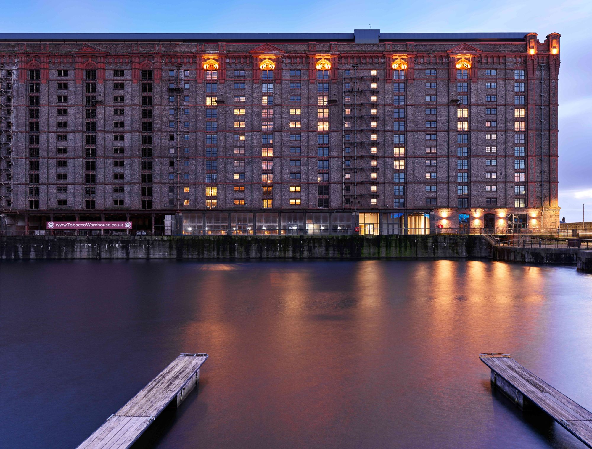 Night time photograph of large brick building by body of water with the lights shining in half of the windows.