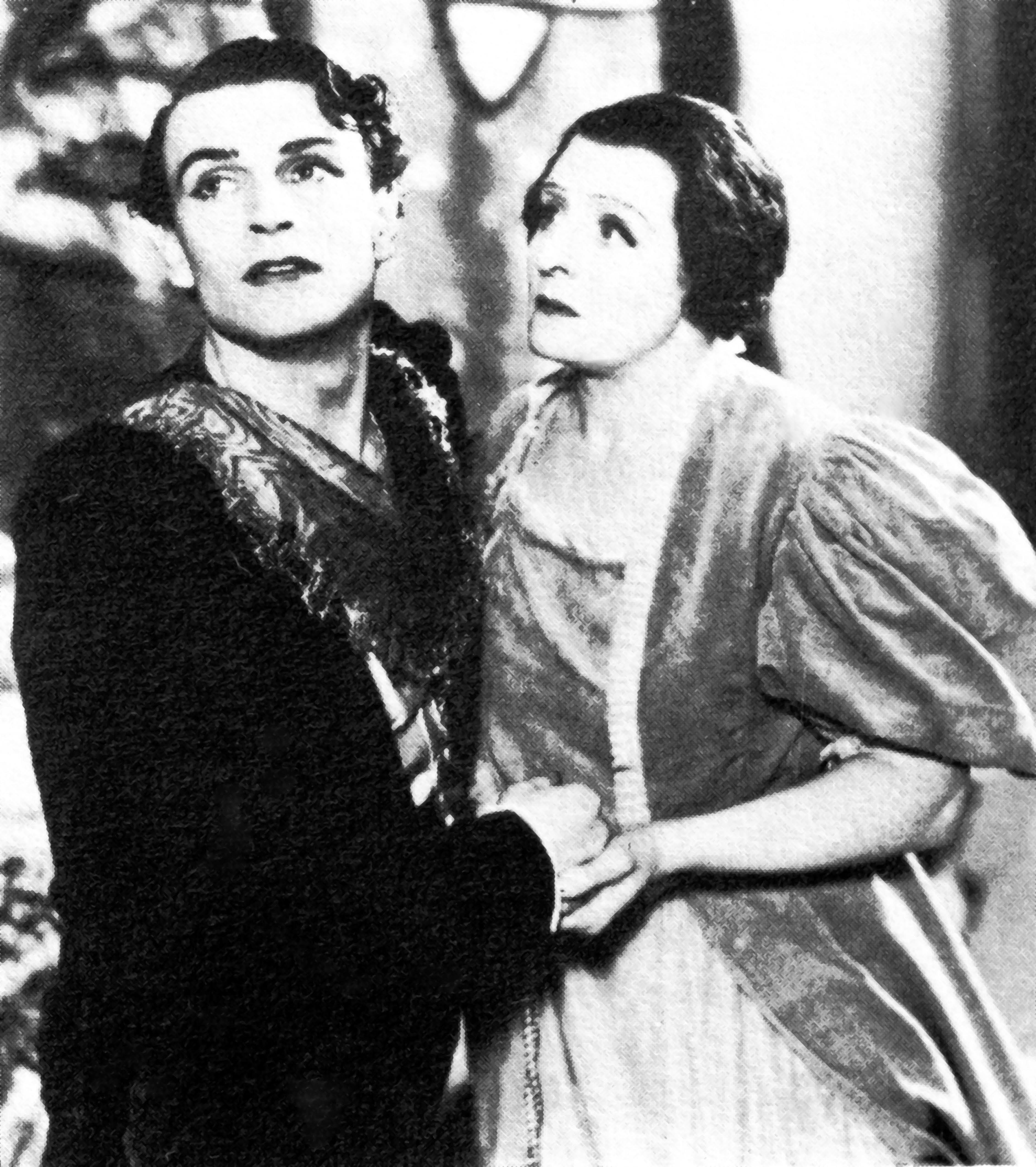 A black and white photo of two actors embracing. The right actor is looking at the left actor, who is looking off into the distance.