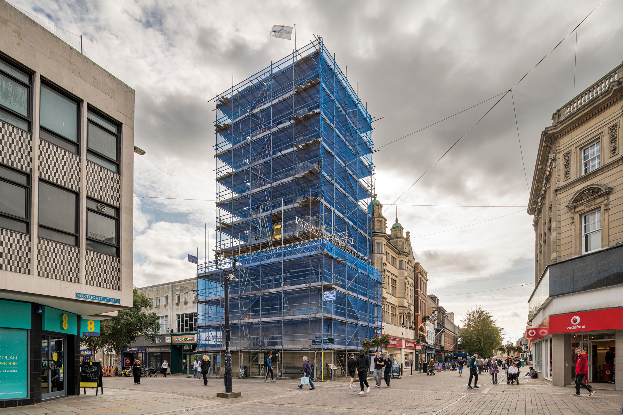A view across a high street crossroads to a medieval church tower covered in scaffolding.