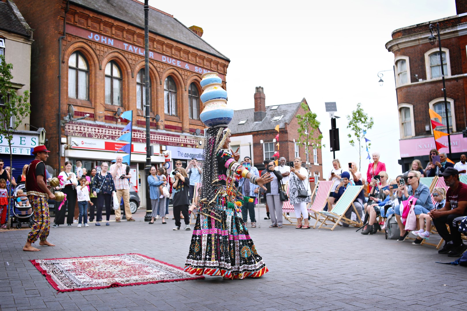 2 dancers in traditional Rajasthan costume dance in front of a crowd of onlookers. In the background are high street shops.