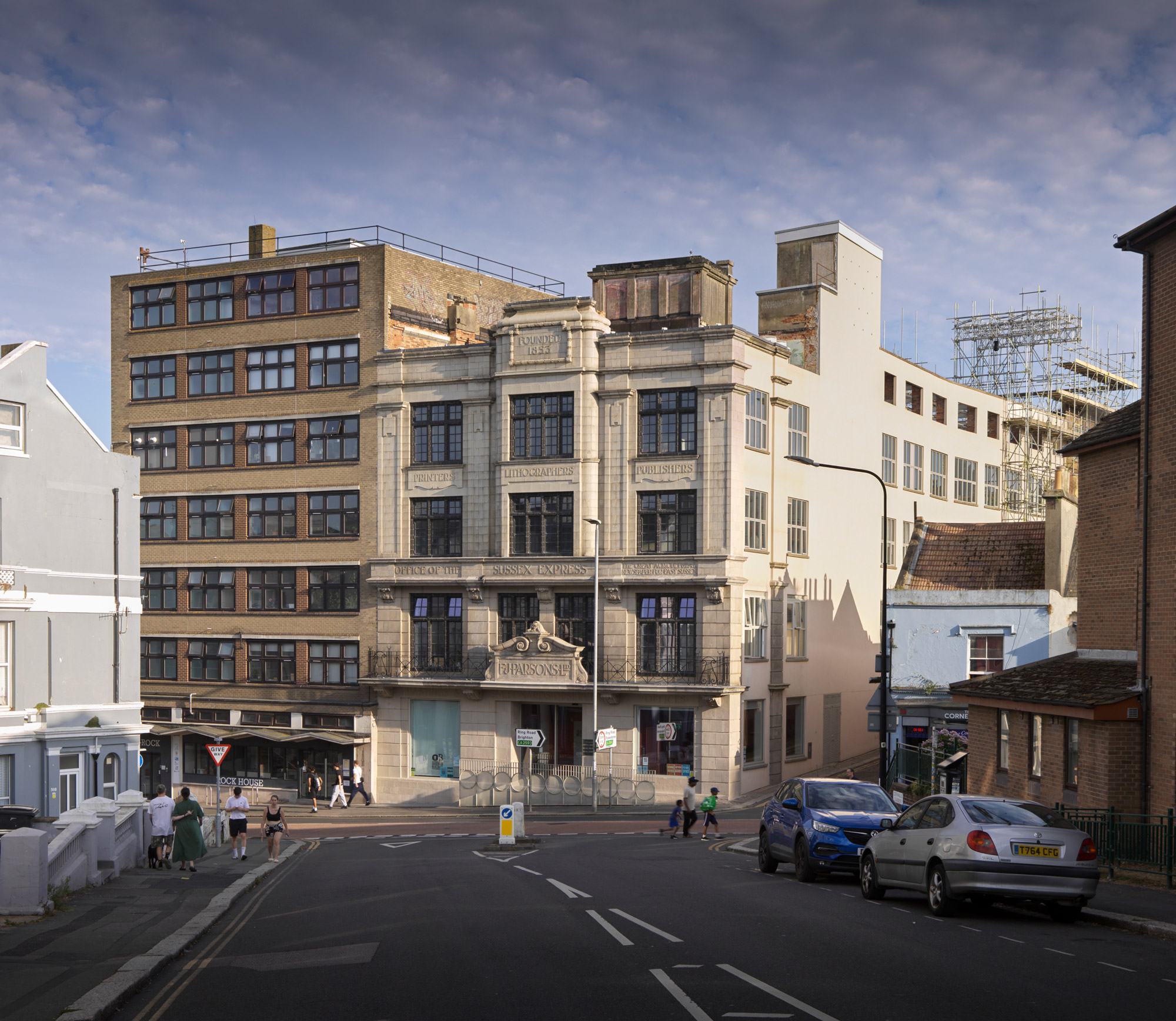 Colour photo of a 4-storey office building with faience and glazed terracotta facade. In the foreground is a quiet street with parked cars and a few pedestrians.