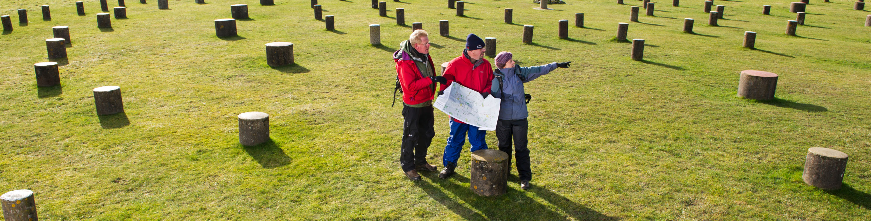 A wide photo of three visitors dressed in coats, hats and walking gear in a grass field surrounded by log stumps. The central visitor is holding a map, with another listening and another pointing off into the distance.
