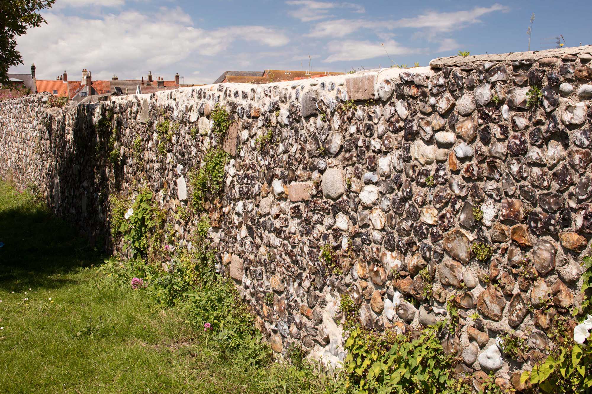 A knapped flint stone wall recedes into the distance. Over the top of it, some clay-tiled roofs are just visible.
