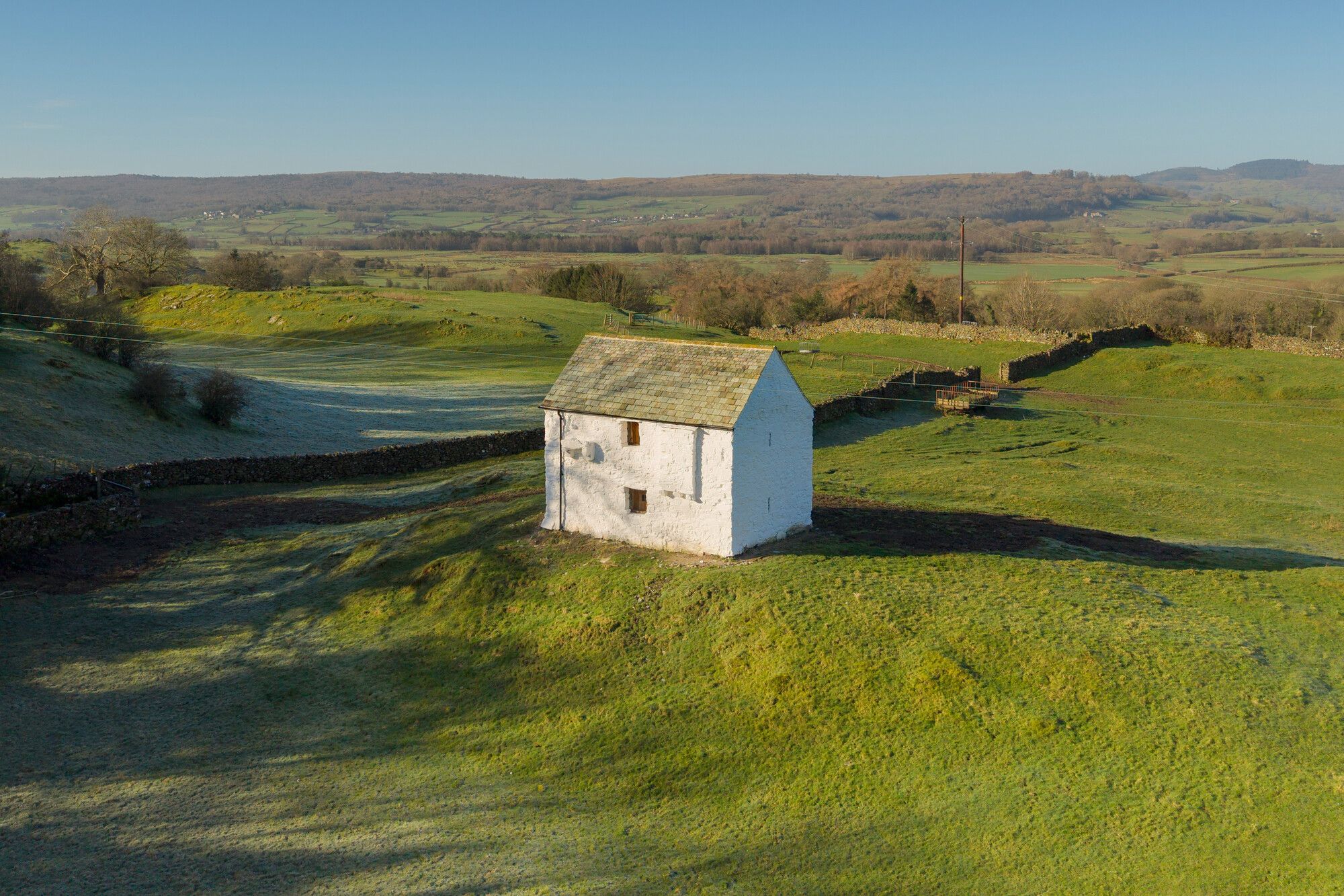 Infront of a forest and field landscape, stands a small, white brick, 2 story building sat on-top of a small grass mound.