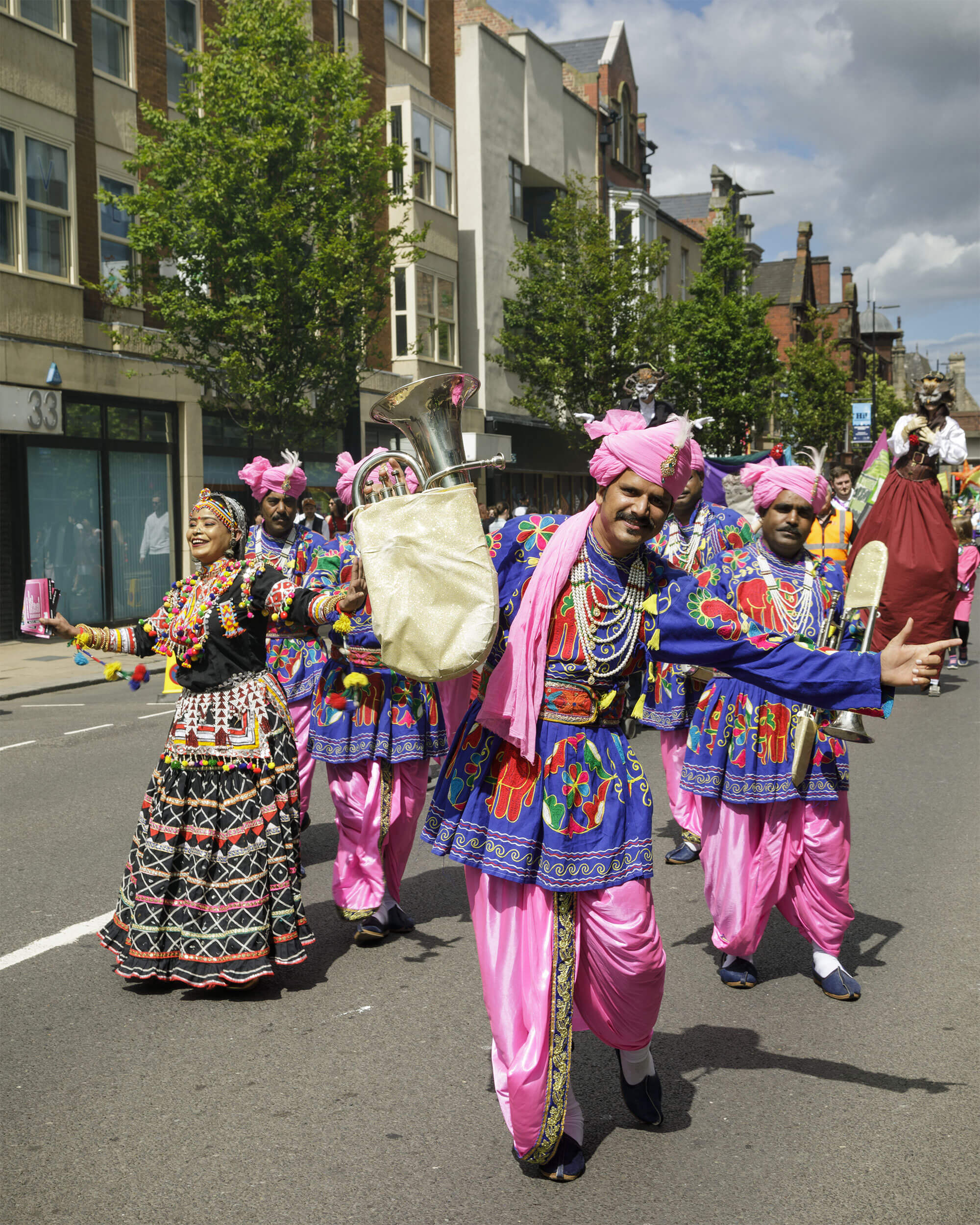 A vibrant street parade featuring performers in colourful traditional attire, adorned with intricate patterns and accessories, celebrating culture with lively expressions and energetic movements under a bright blue sky.