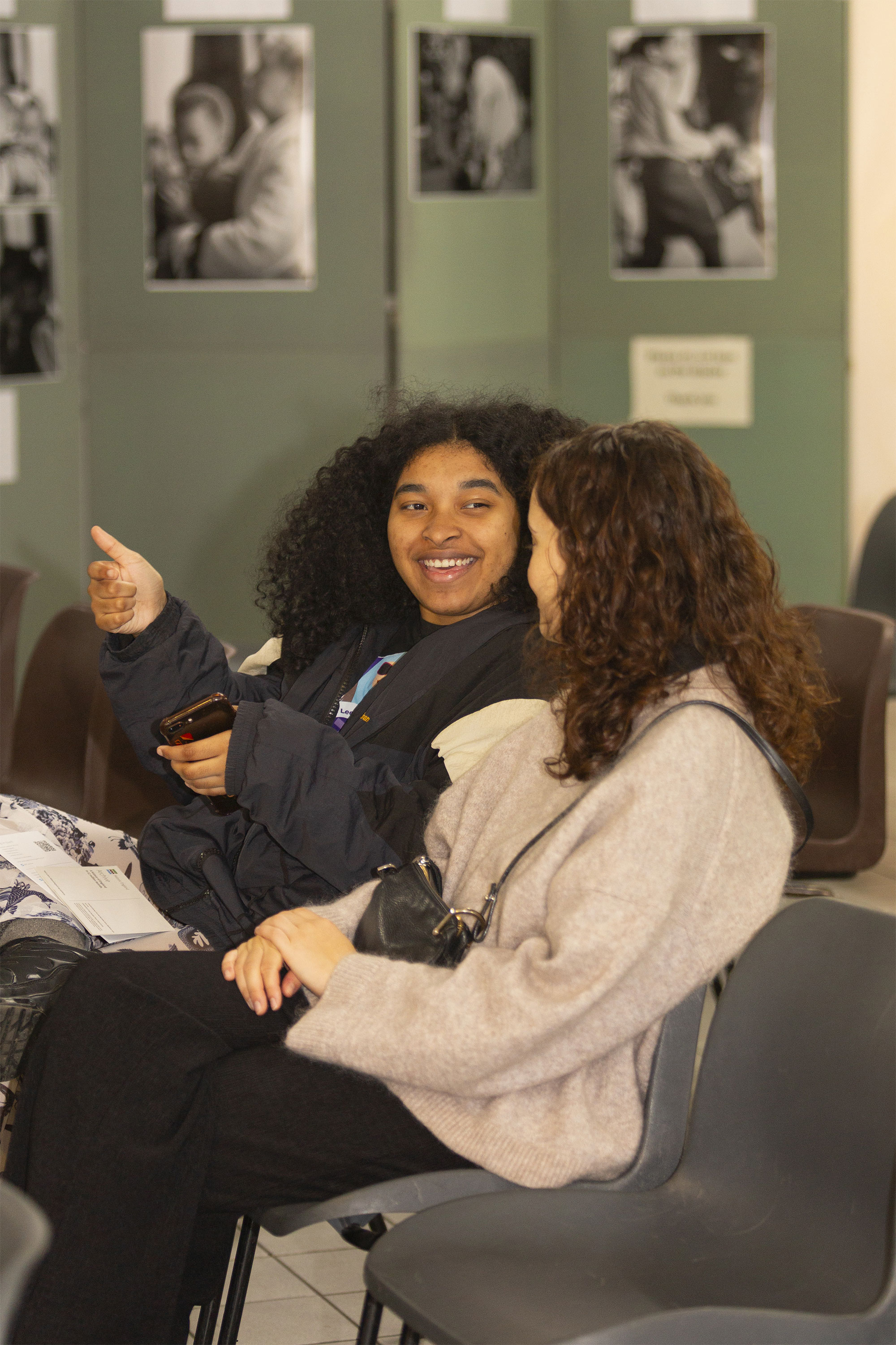 Two young women are seated in a casual setting, engaged in conversation. One has curly hair and is smiling, while the other listens attentively, creating a friendly atmosphere.