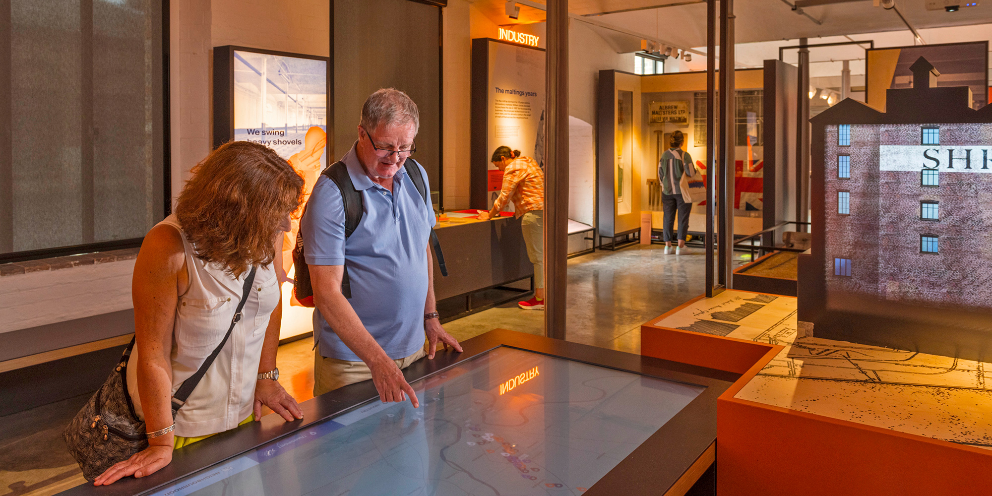 A photo of an interior exhibition space, with two people looking at a screen in the foreground and others looking at other exhibits in the background