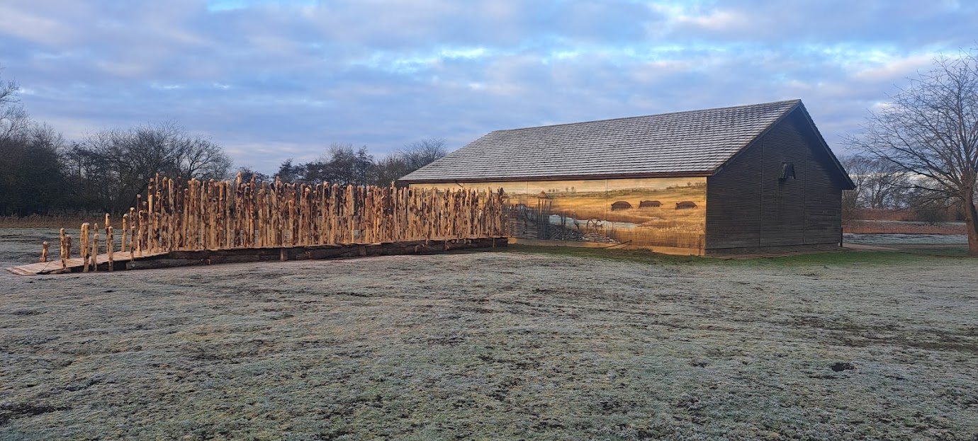 The photo shows the created Bronze Age timber walkway with upright posts and timber flooring, and the mural on the side of the building at the end of the walkway with water, reeds and landscape in the distance 