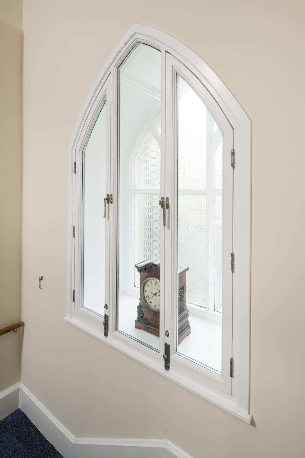 An arched window with 3 panes of secondary glazing in front of the original window. A wooden clock sits on the window cill. 