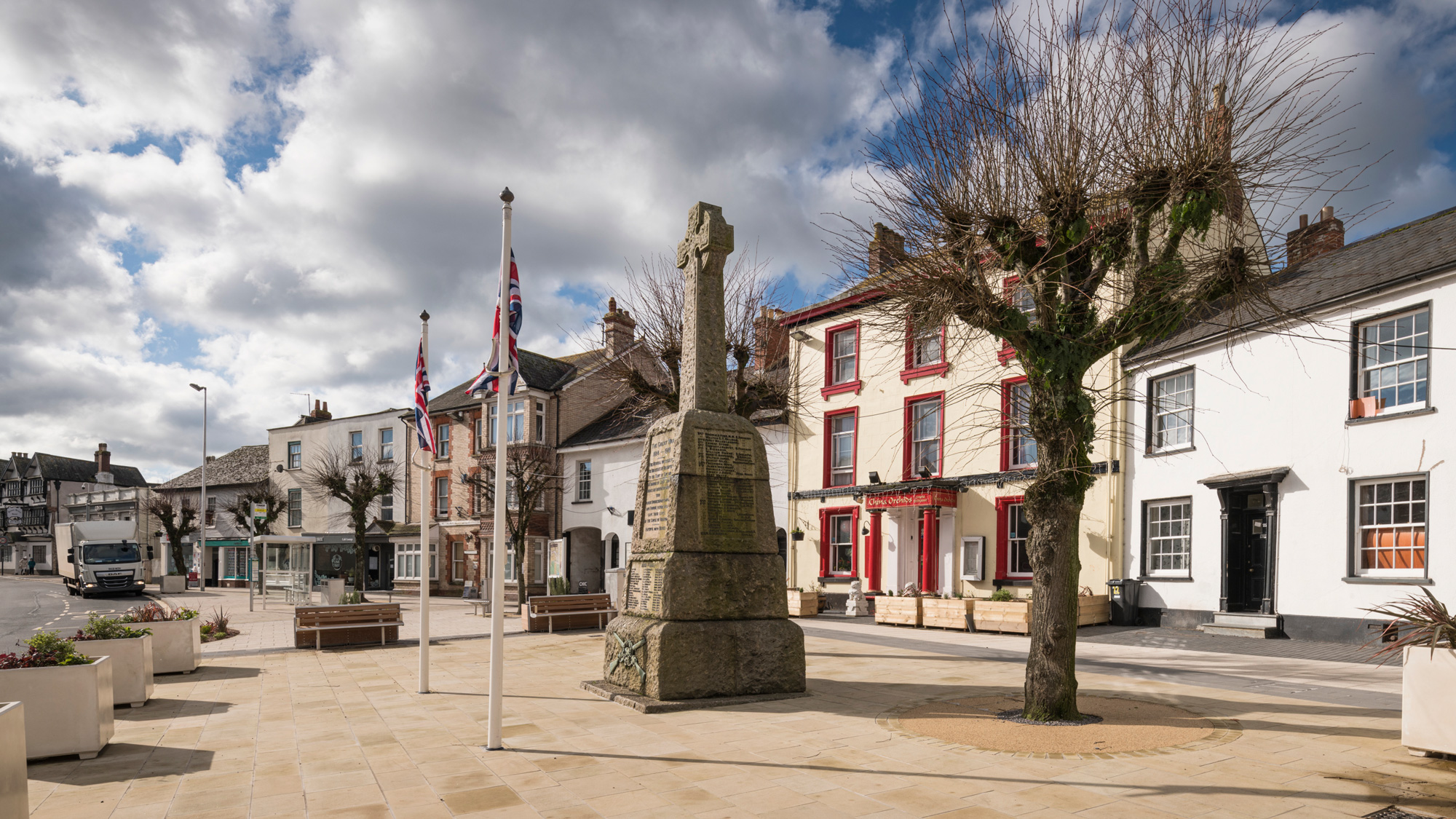 War memorial on a street of older buildings. The memorial is surrounded by newly laid flagstones, planting and 2 flagpoles