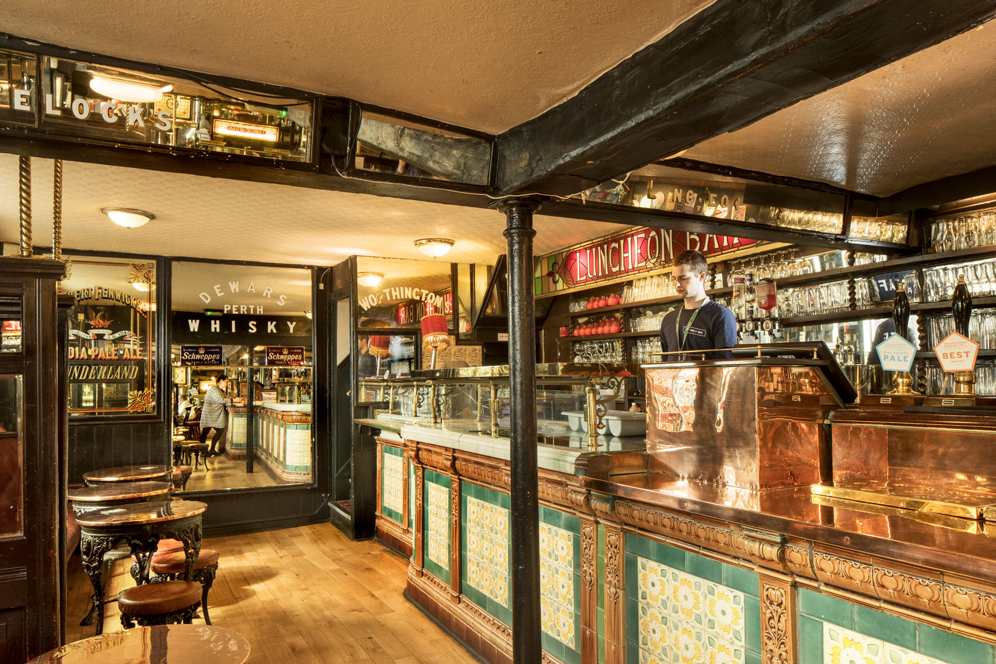 Bar with tiled front and sign behind saying 'LUNCHEON BAR'. On a glass door and mirrored walls there's advertising for various drink brands.