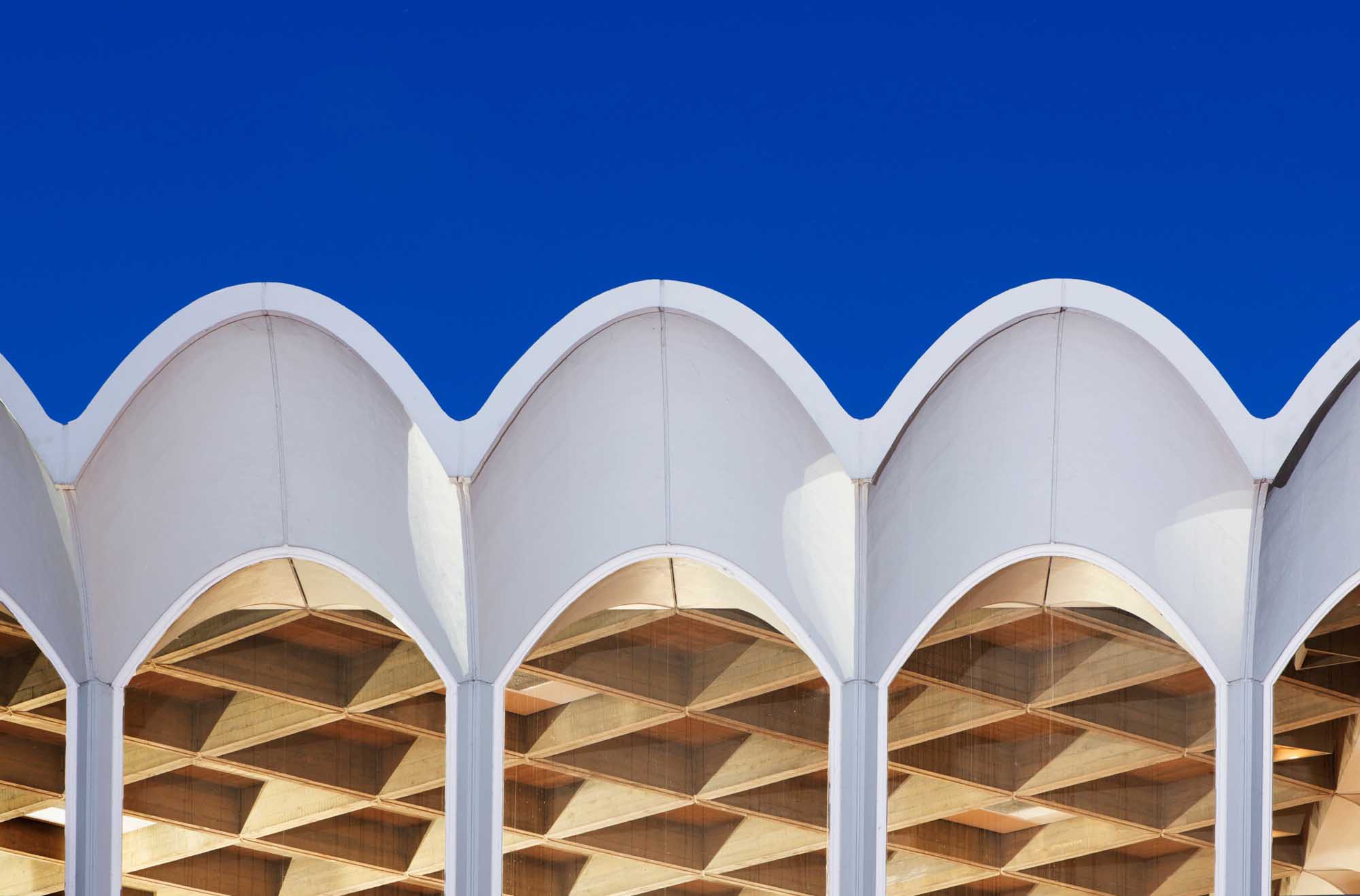 A close up image of the west elevation of The Central Hall shows white archways against a bright blue sky, with a gridded roof behind.