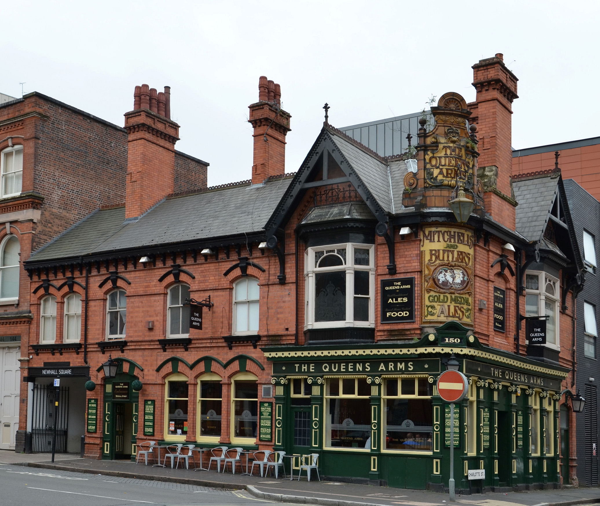 A street-level photo of a 2-storey red brick public house with green and gold frontage reading "The Queen's Arms"