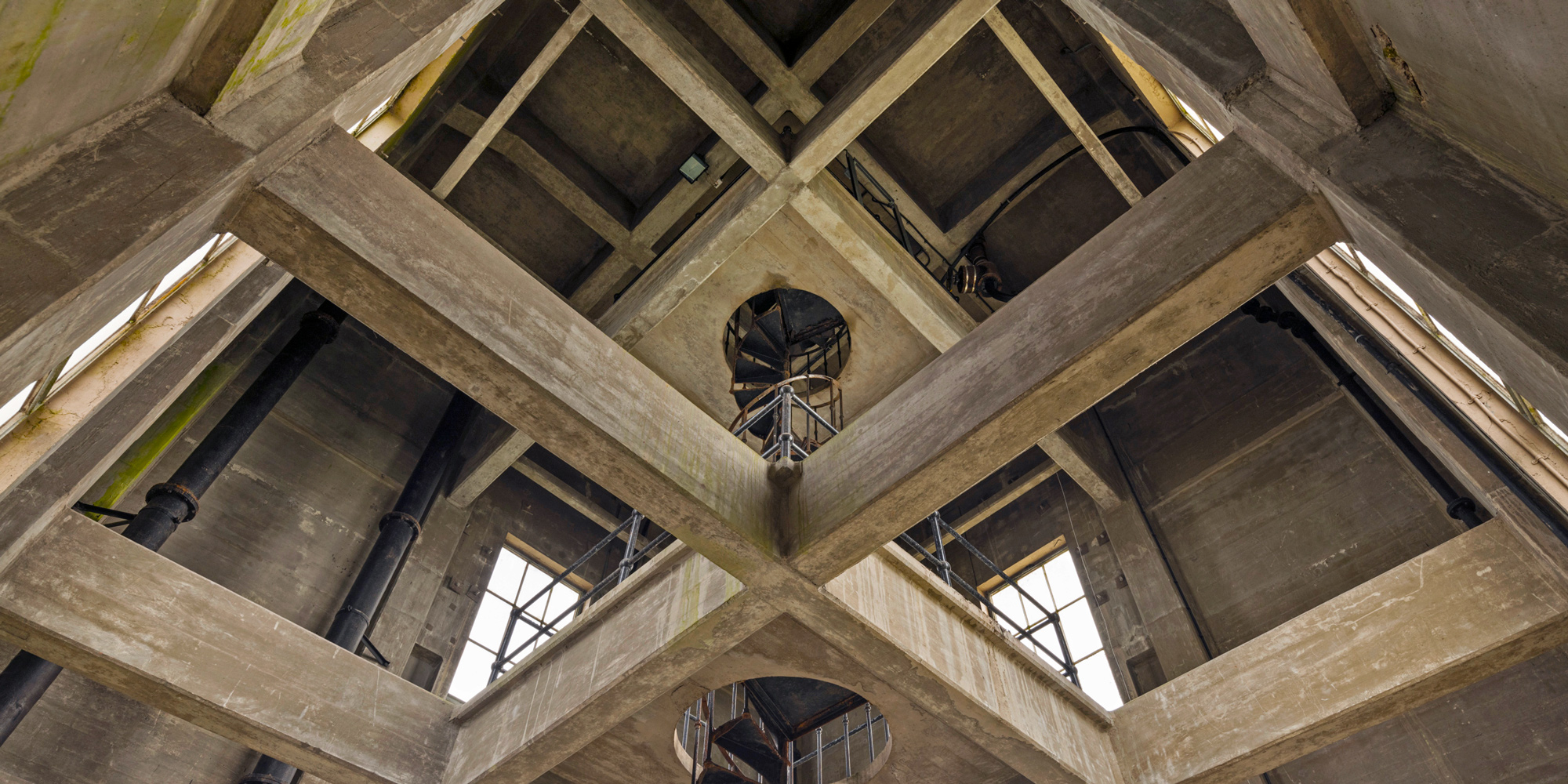 A spiral staircase in the centre of a large concrete building cross-braced with concrete trusses. 
