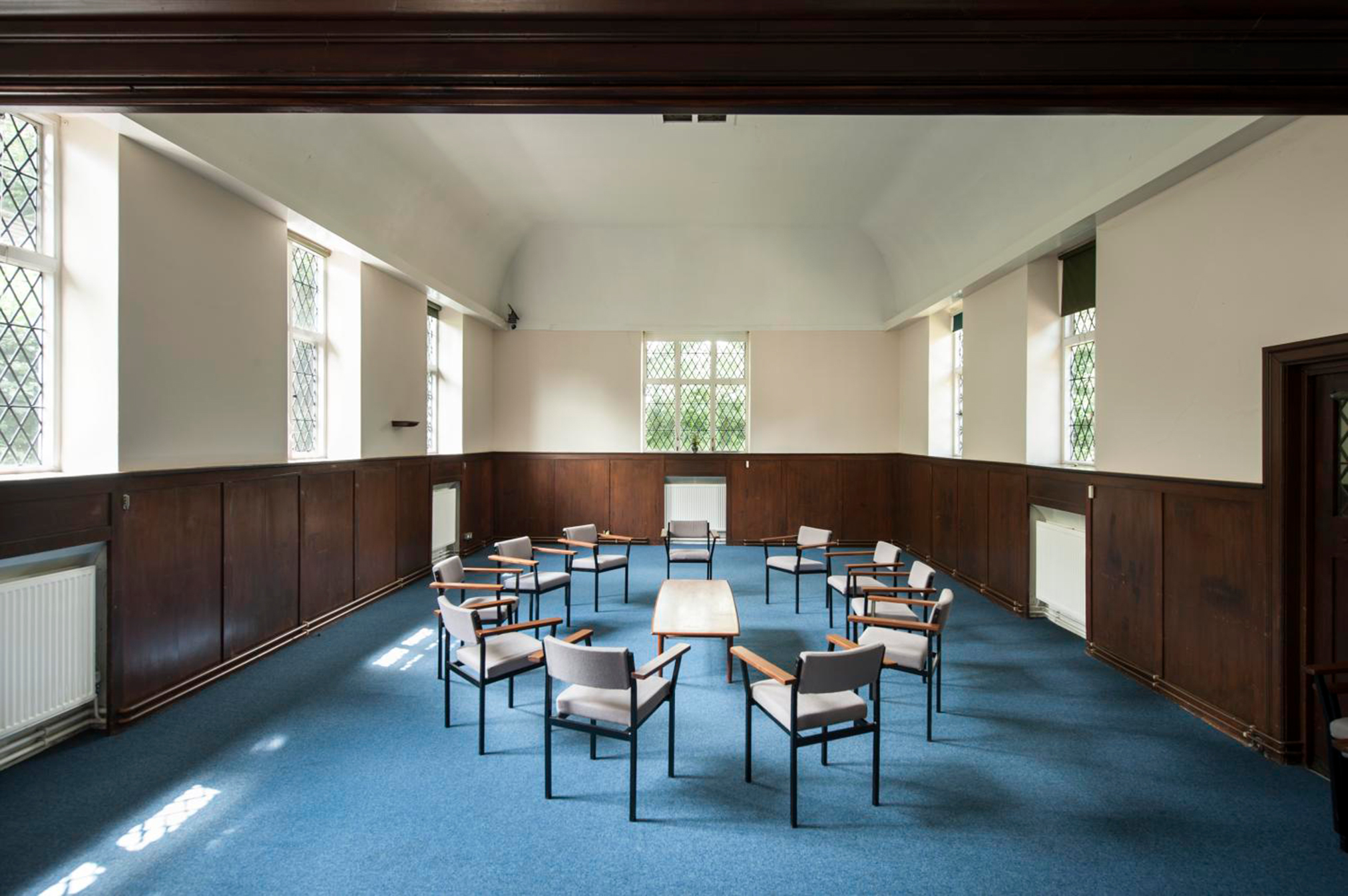 Interior of a meeting room with tables and chairs arranged centrally, surrounded by large windows and wood paneling.