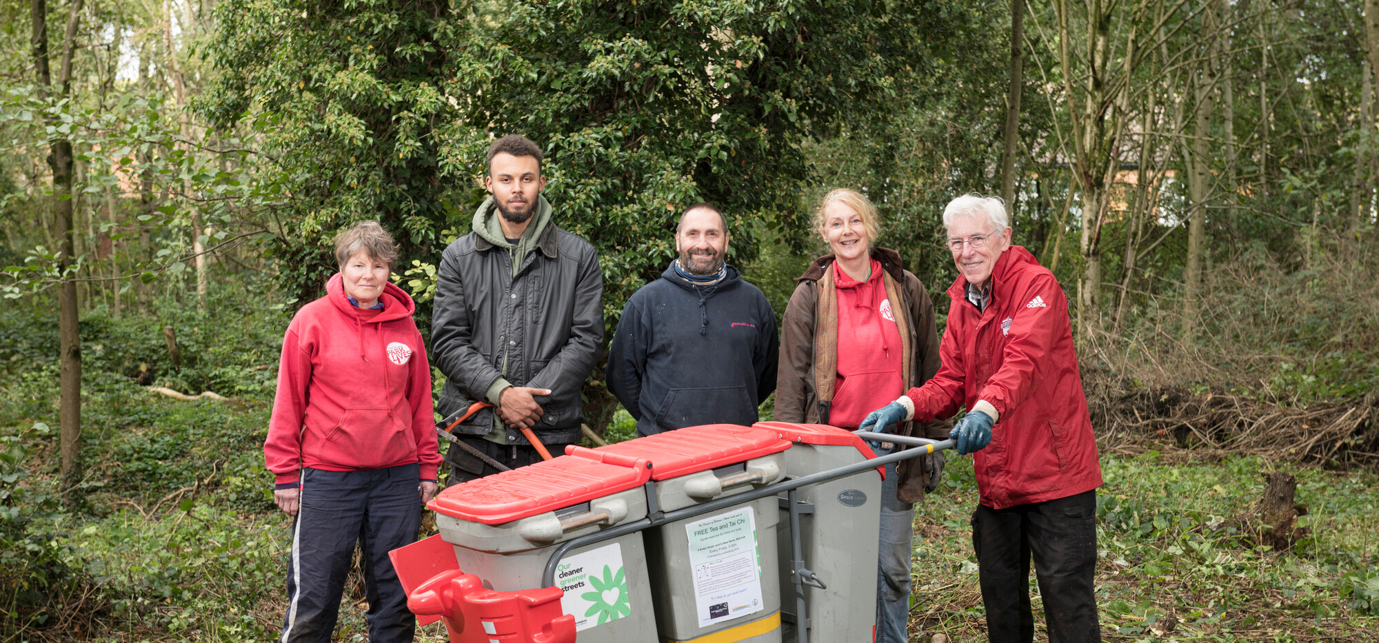 A group of 5 people stands in a wooded area, smiling beside a recycling cart. They are dressed in casual outdoor clothing, ready for a community clean-up effort.