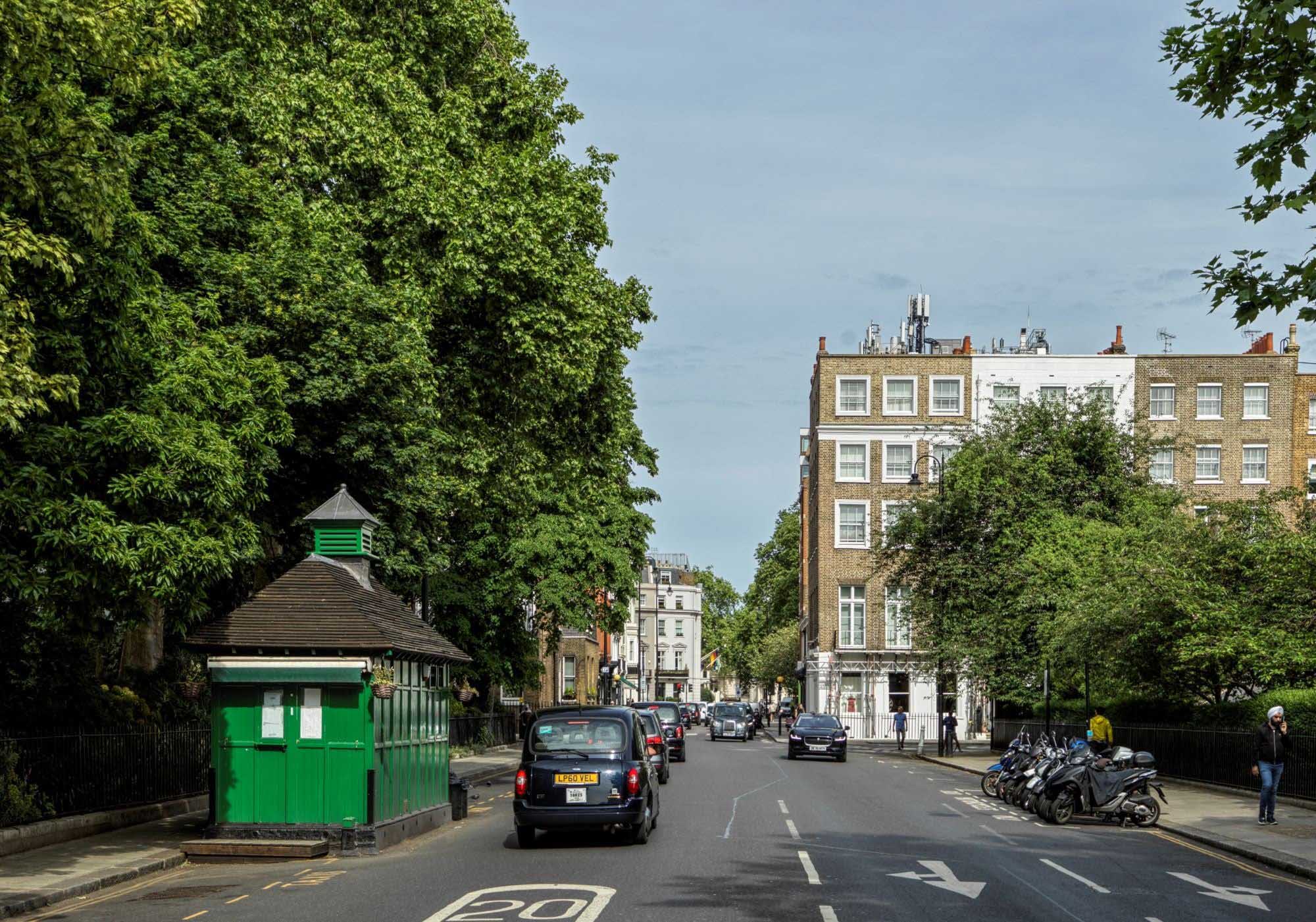 A small green wooden hut with a pointed roof next to a road with a line of cars driving past