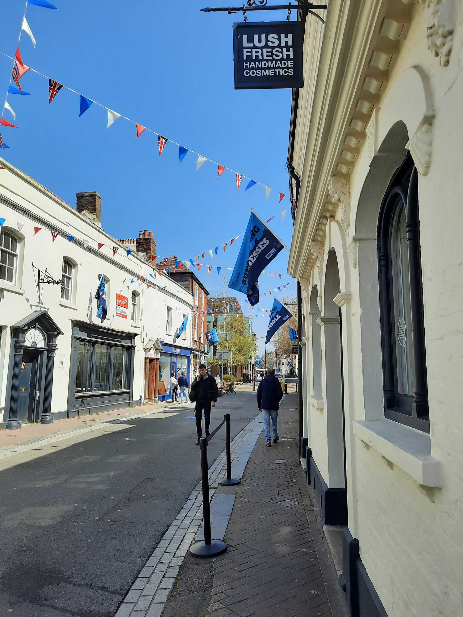 A man walking in a high street lined with historic shops. There is coloured bunting strung across the street.