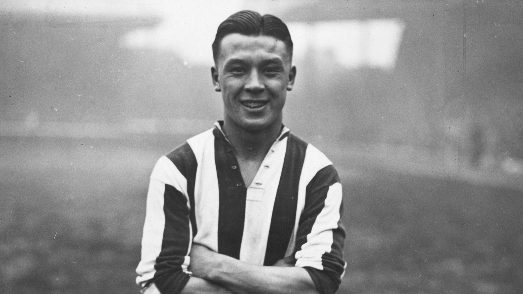 A black and white photograph of a footballer with his arms crossed wearing a striped football shirt.