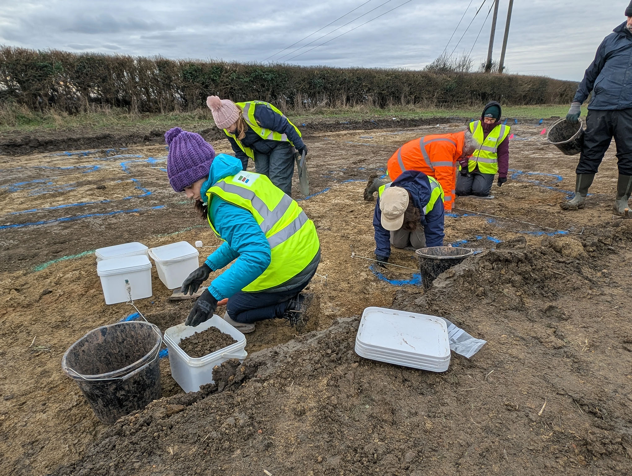 Historic England and community volunteers on their knees in a muddy trench from the Willersey dig. 