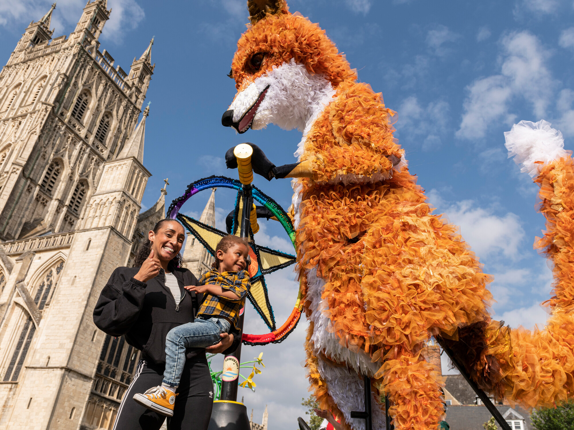 A woman holds a child while a large, colorful fox puppet stands beside them, set against a backdrop of a historic building and a bright blue sky.