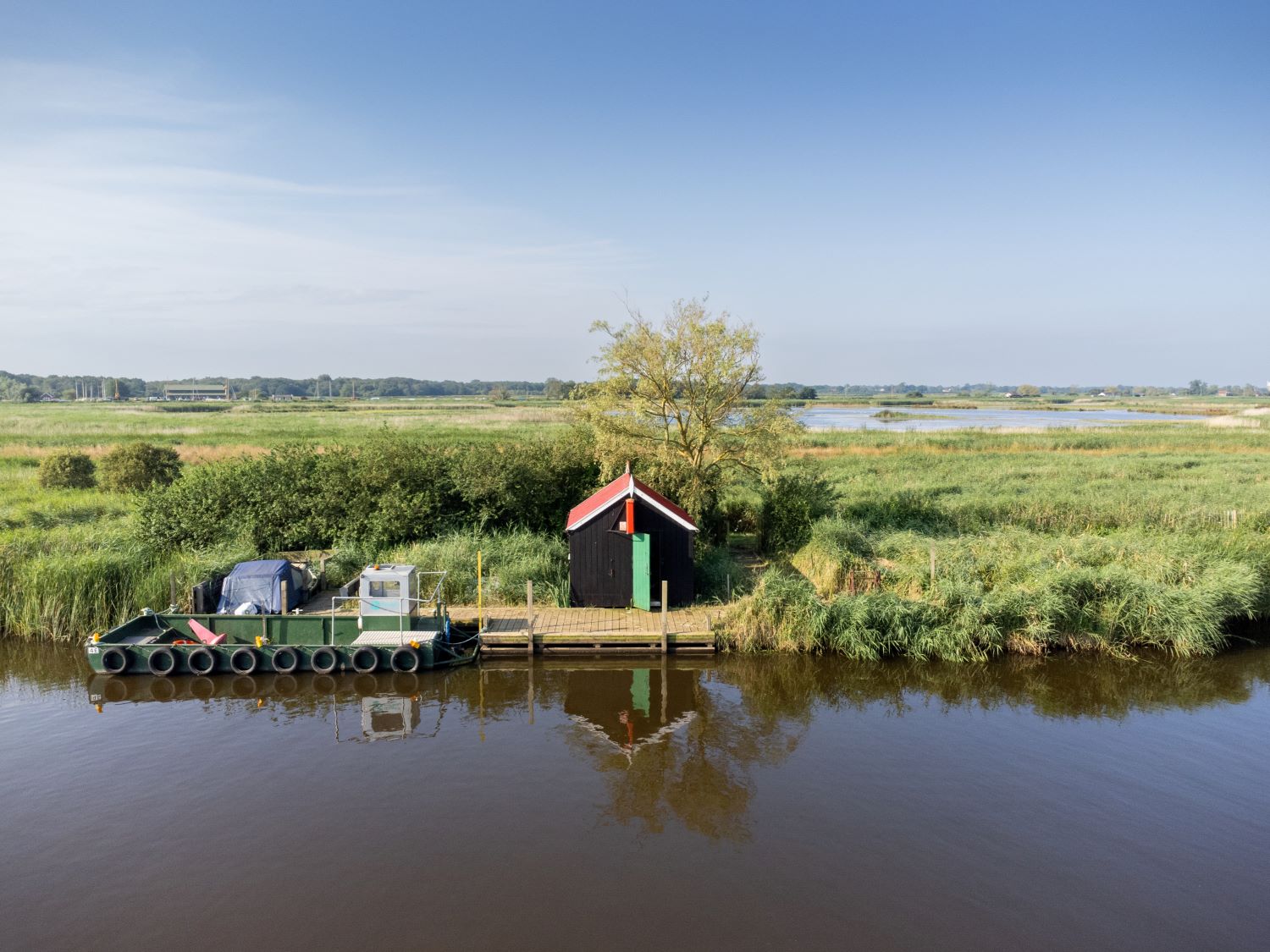 The photograph shows the river in the foreground with a small brown shed-like building on a platform with green door and red roof, with a red Eel Stop sign. The wider Broads National Park landscape can be seen in the background.