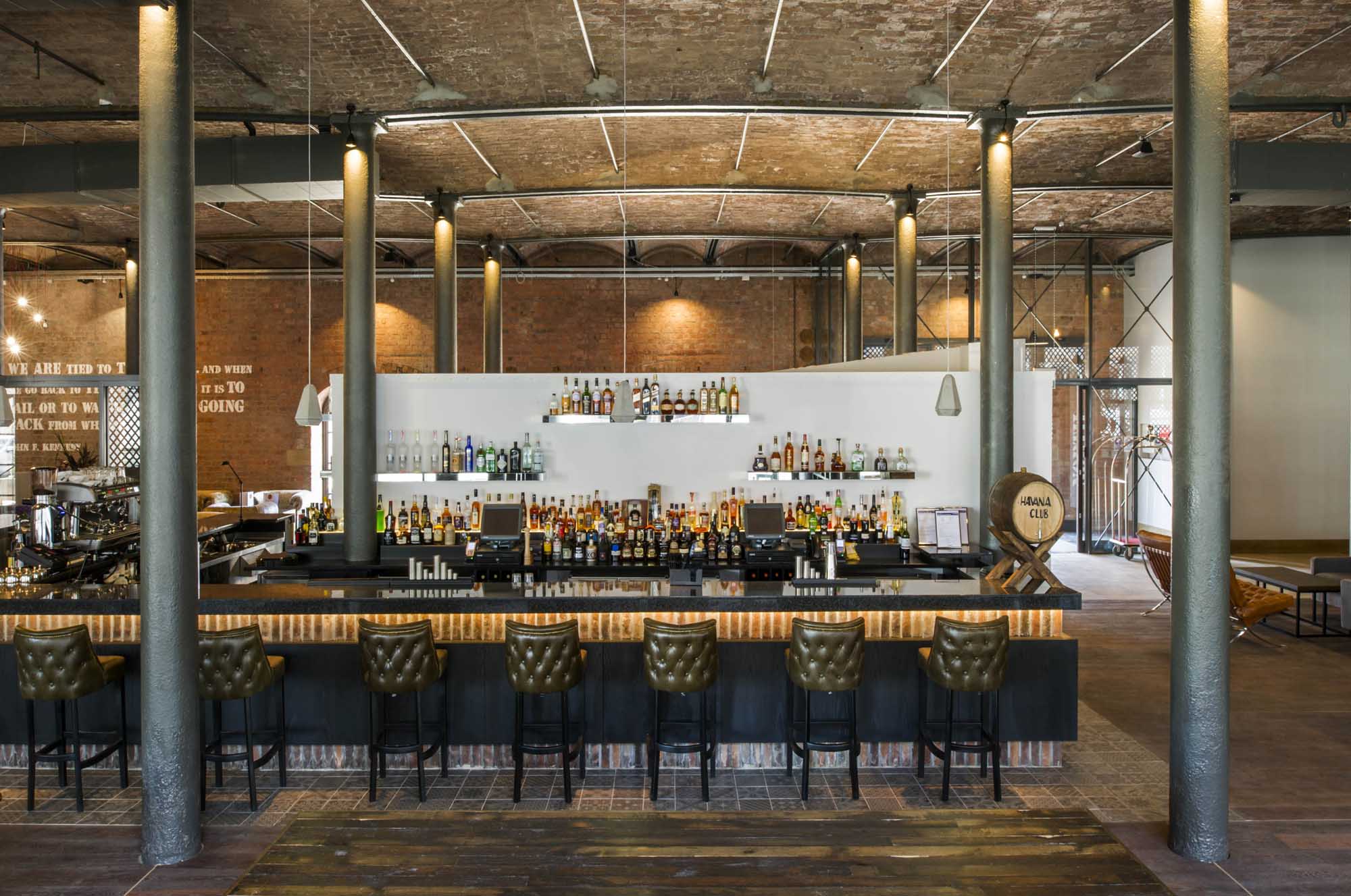 Chairs lined up at a bar in a high-ceilinged room with iron pillars and a red brick wall.