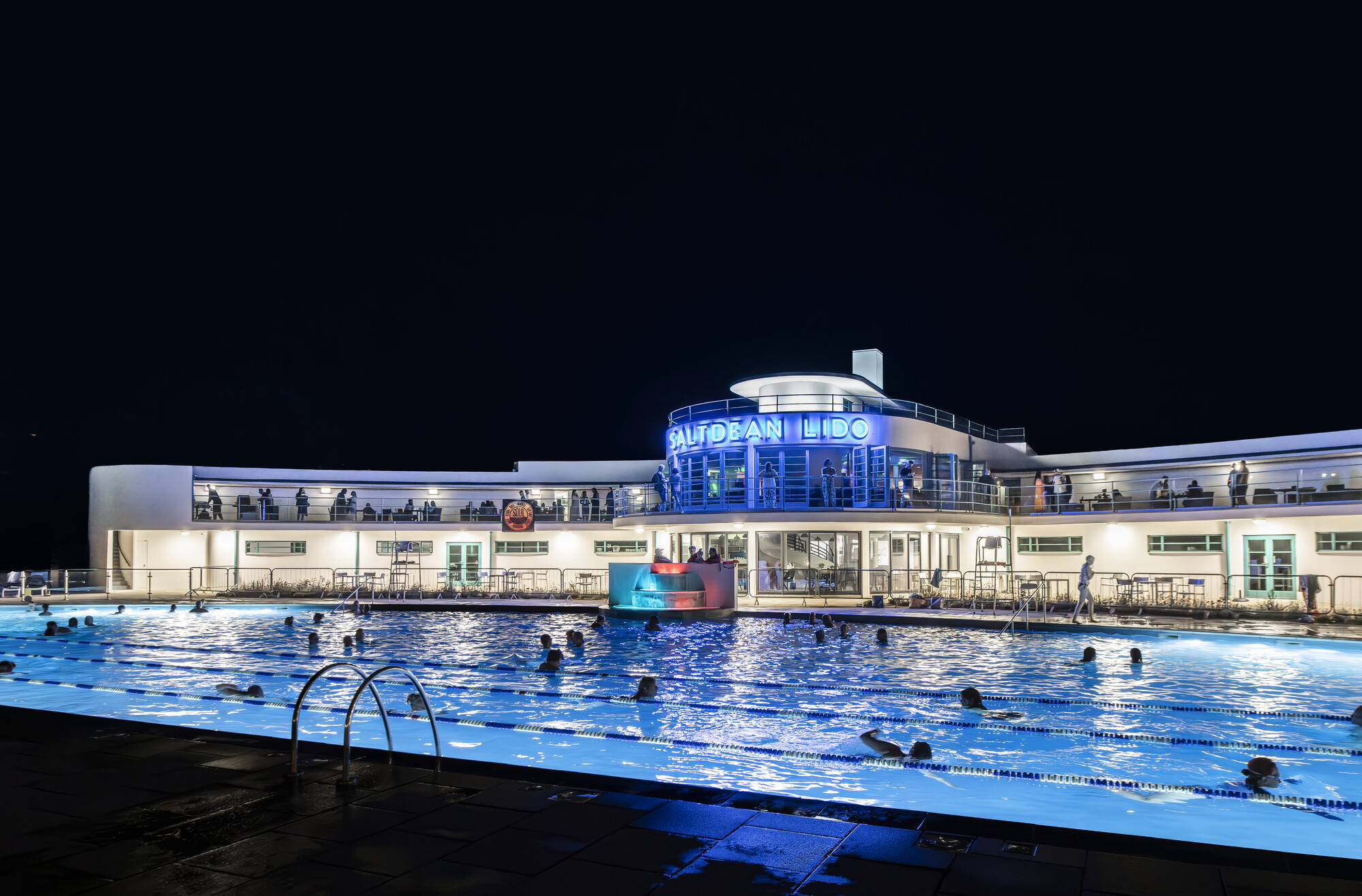 An art deco lido at night, featuring a brightly lit pool surrounded by people. The building's sleek design and vibrant lighting create a lively atmosphere for evening swimming and relaxation.