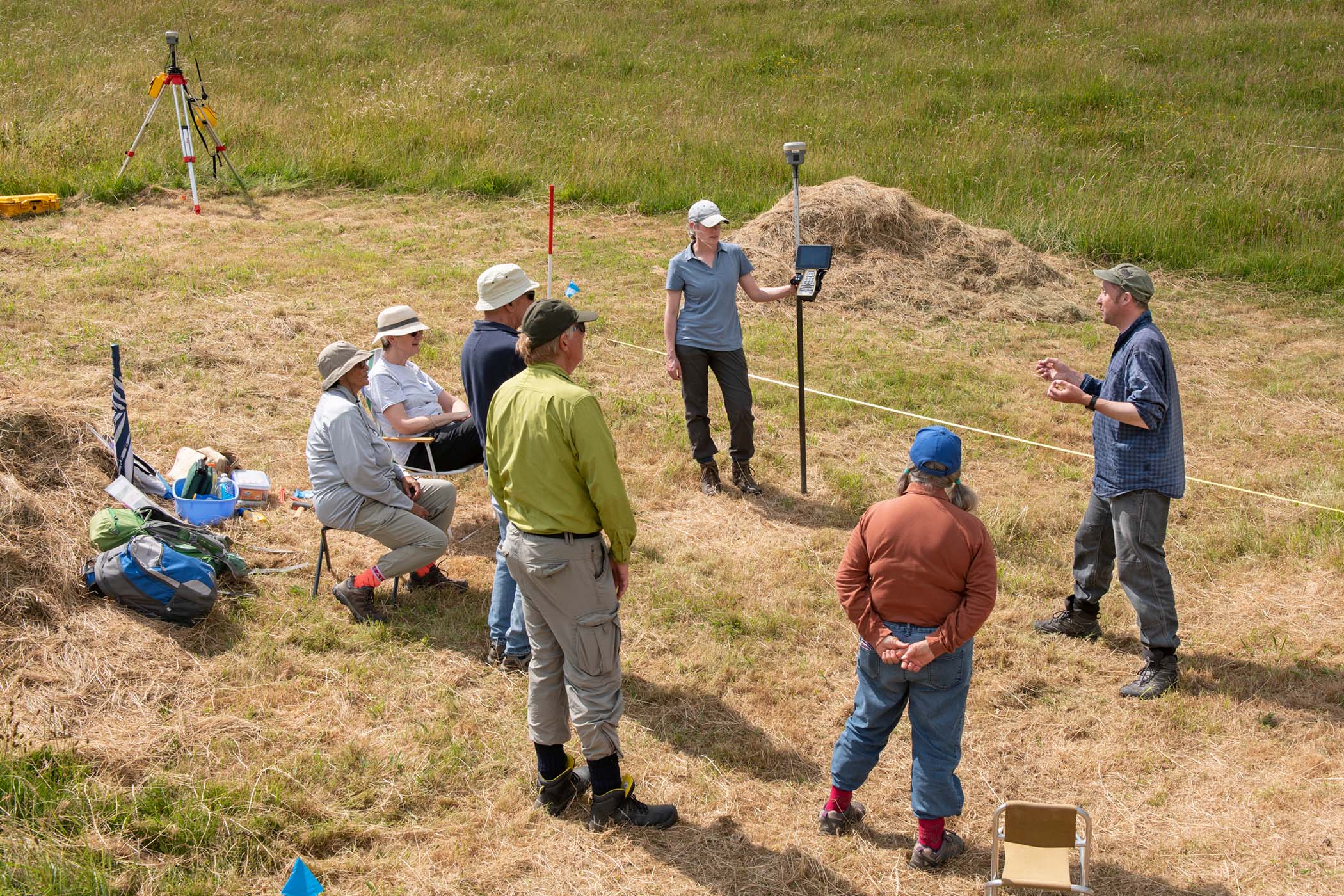 A group of people, some seated and some standing, gather in a grassy field around a person holding a survey pole and tablet. Surveying equipment and measuring tapes are visible.