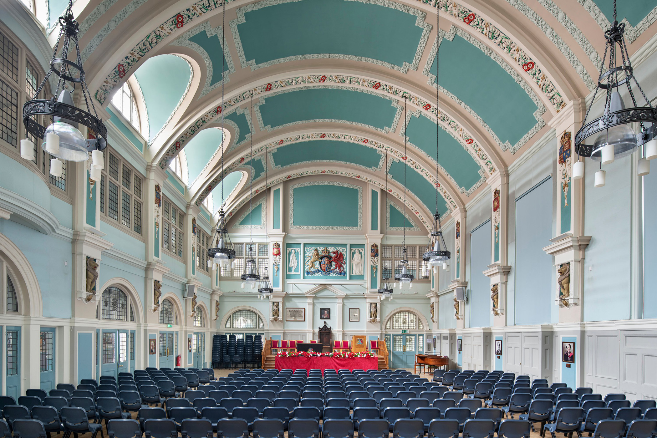 Interior wide-angle photograph of a school hall with an ornate ceiling above and rows of blue chairs in the foreground