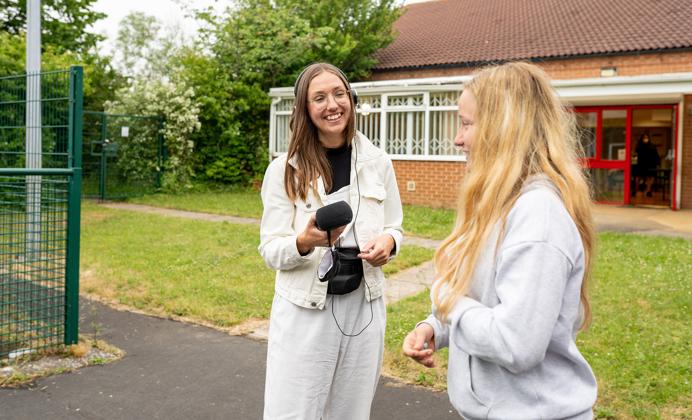A person with long hair and round glasses dressed in white trousers and jacket holding a microphone and wearing headphones smiles at a person with long blonde hear wearing a grey sweatshirt outside a single storey brick building and grassy area.