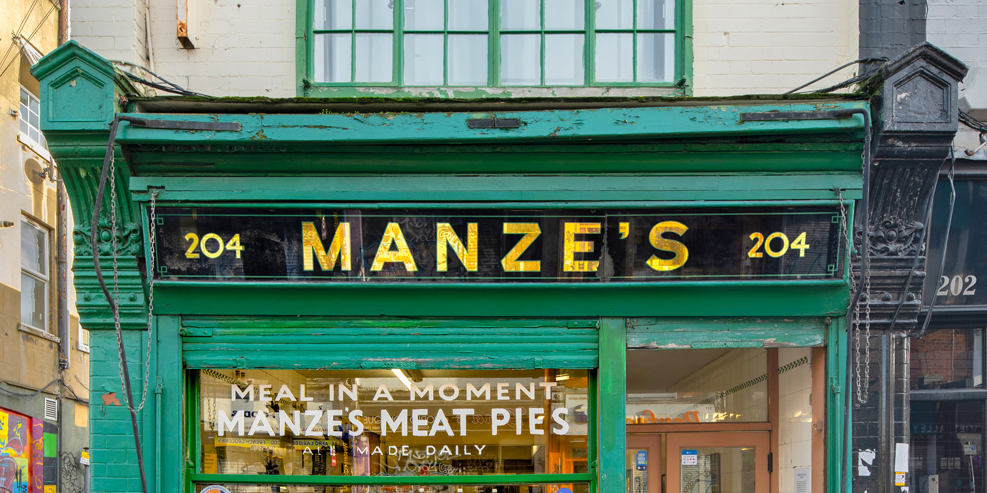 A green shop front with a sign saying "204, Manze's, 204; Meal in a moment, Manze's Meat Pies, all made daily".