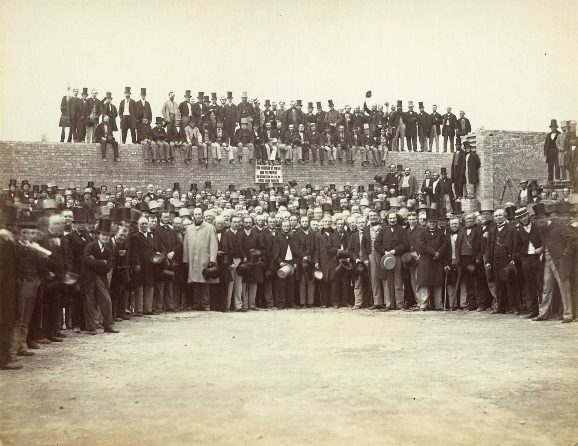 A large group of men posed in front of and on top of the brick wall forming part of the overflow reservoir at Crossness Pumping Station.  Most of the men are smartly dressed complete with top hats. A sign on the wall reads “WEIR WALLS FOR OVERFLOW OF SEWAGE AND TO PREVENT RESERVOIR BEING UNDER PRESSURE”.