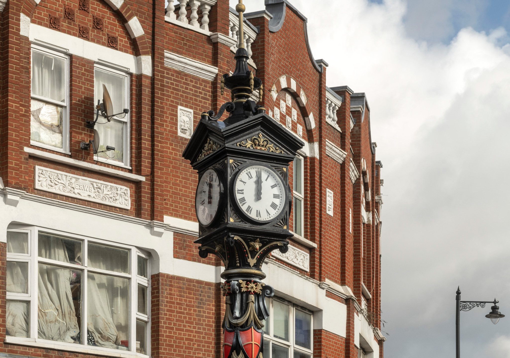 A clock set in an ornate iron housing stands above a high street and shown here with period buildings behind and without ornate iron base at pavement level cropped out. 2 out of the clock's 4 faces are visible in this view.
