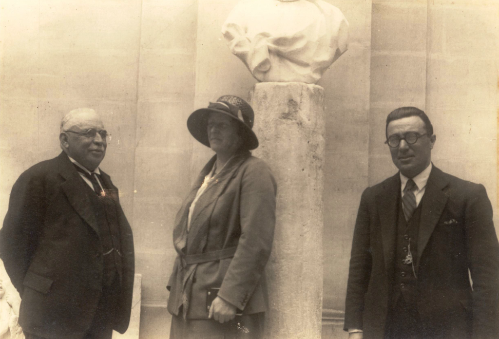 A sepia photograph of a woman stood between 2 men in front of a marble bust mounted on a stone column pedestal.