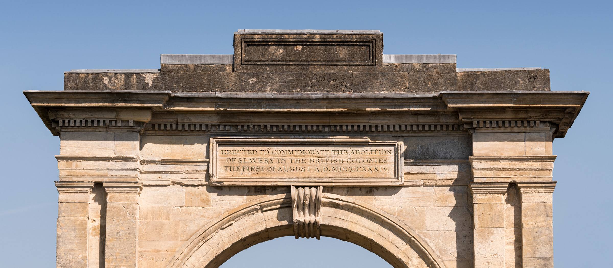 Photograph of a stone arch between the arch and cornice on the east side. A rectangular plaque reads: 'Erected to commemorate the abolition / of slavery in the British colonies / The first of August A.D. MDCCCXXXIV.'