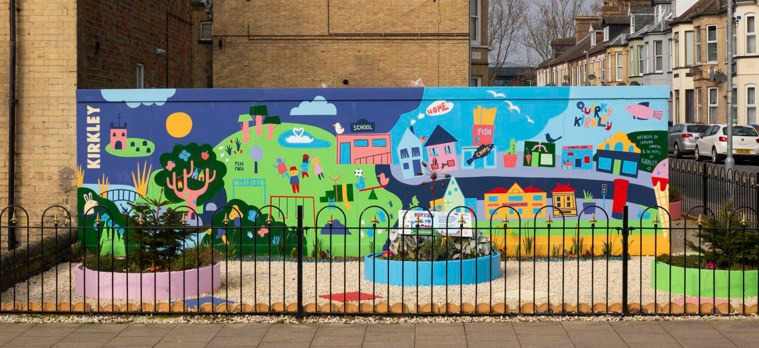 A wall with bright cartoon-like artwork applied to it which depicts the local place. In front of the wall are three circular planters.