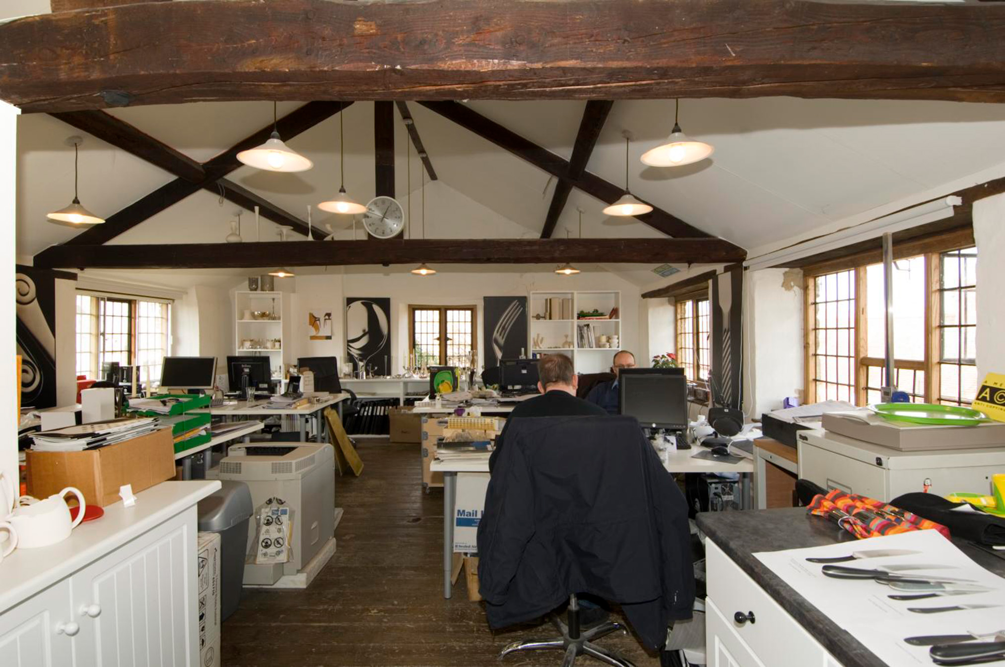 Two people are working at computer desks in an office with dark wooden beams supporting the building. There is cutlery laid on on a table in the foreground and large images of cutlery on the walls.
