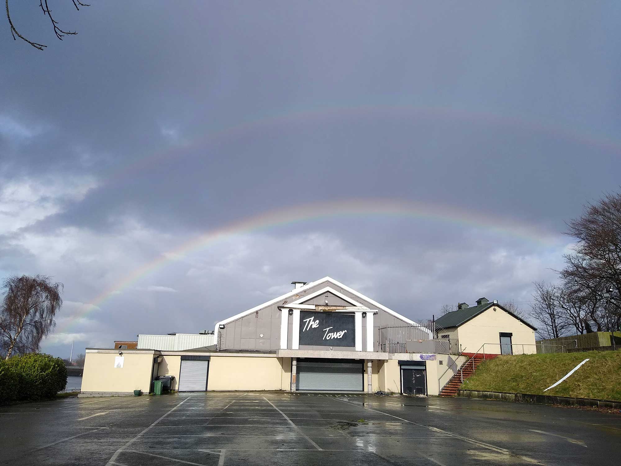 A photo of a 2-storey arch-roof building underneath a stormy sky with a double rainbow. A sign on the front of the building reads: "The Tower".