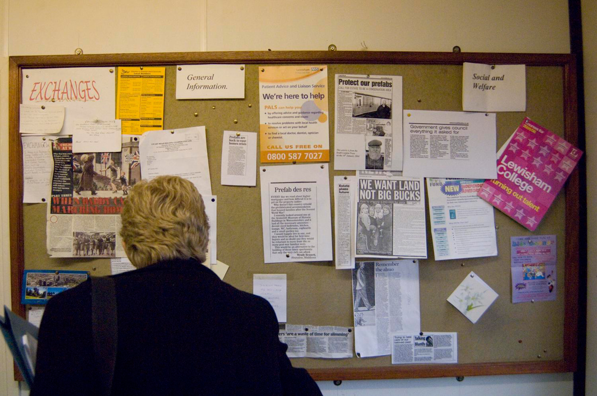A photograph from behind of a person with short wavy hair looking at a General Information noticeboard with newspaper clippings and flyers
