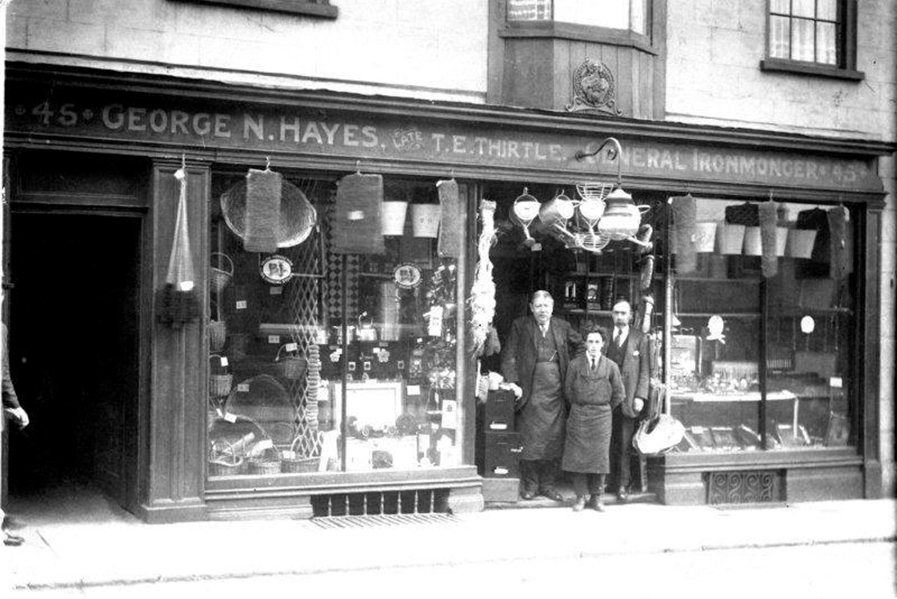 A black and white photograph shows three men wearing shirt, tie, and apron, standing in the doorway of an ironmonger's shop. The large windows either side of the doorway are filled with wares and a tin bath, watering cans and other objects hang above the door.