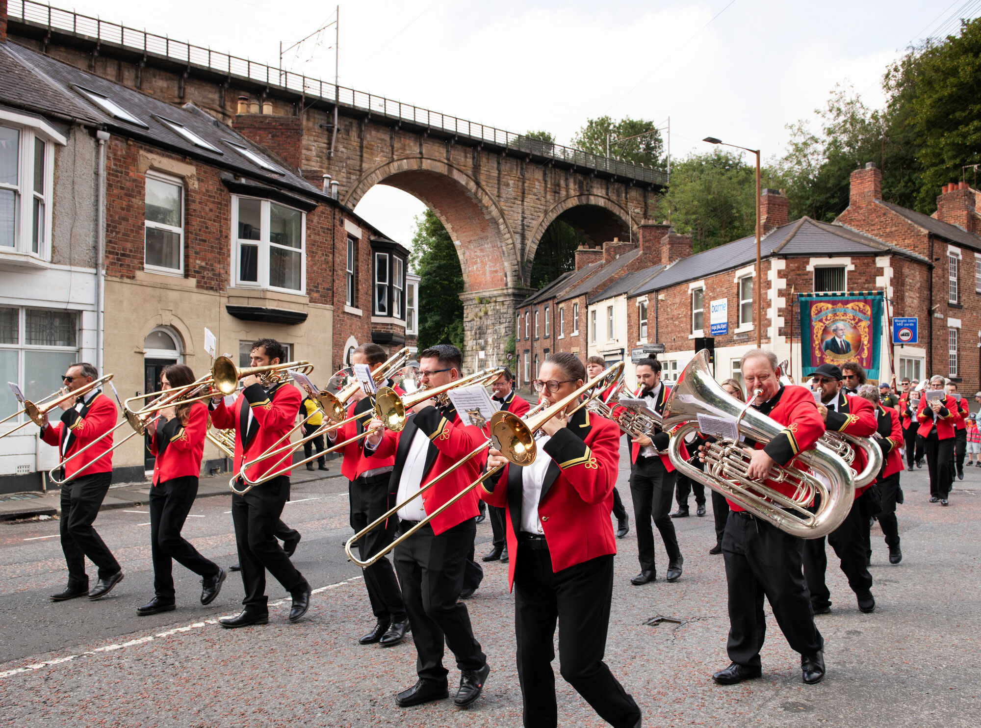 A brass band marches down a street beneath an impressive viaduct, playing for a crowd.