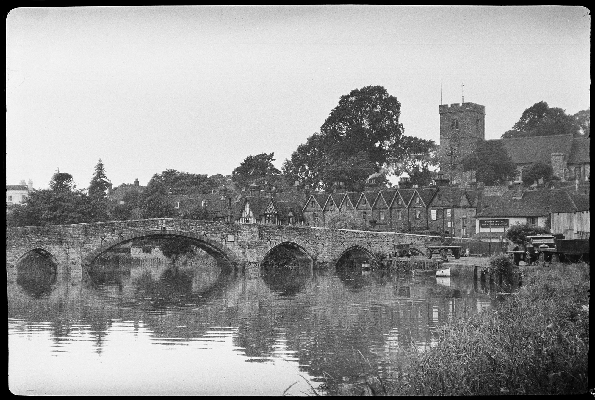 Black and white photograph of a stone bridge spanning a river. Beyond the far bank is a row of houses and an industrial building. Beyond, in an elevated position, is a church with a square tower.