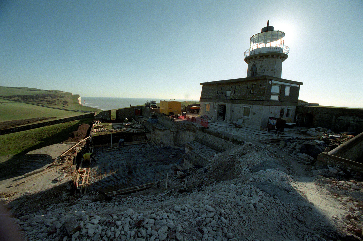 A lighthouse with a building site to the left of it. Clifftops and the sea are visible in the top left corner.