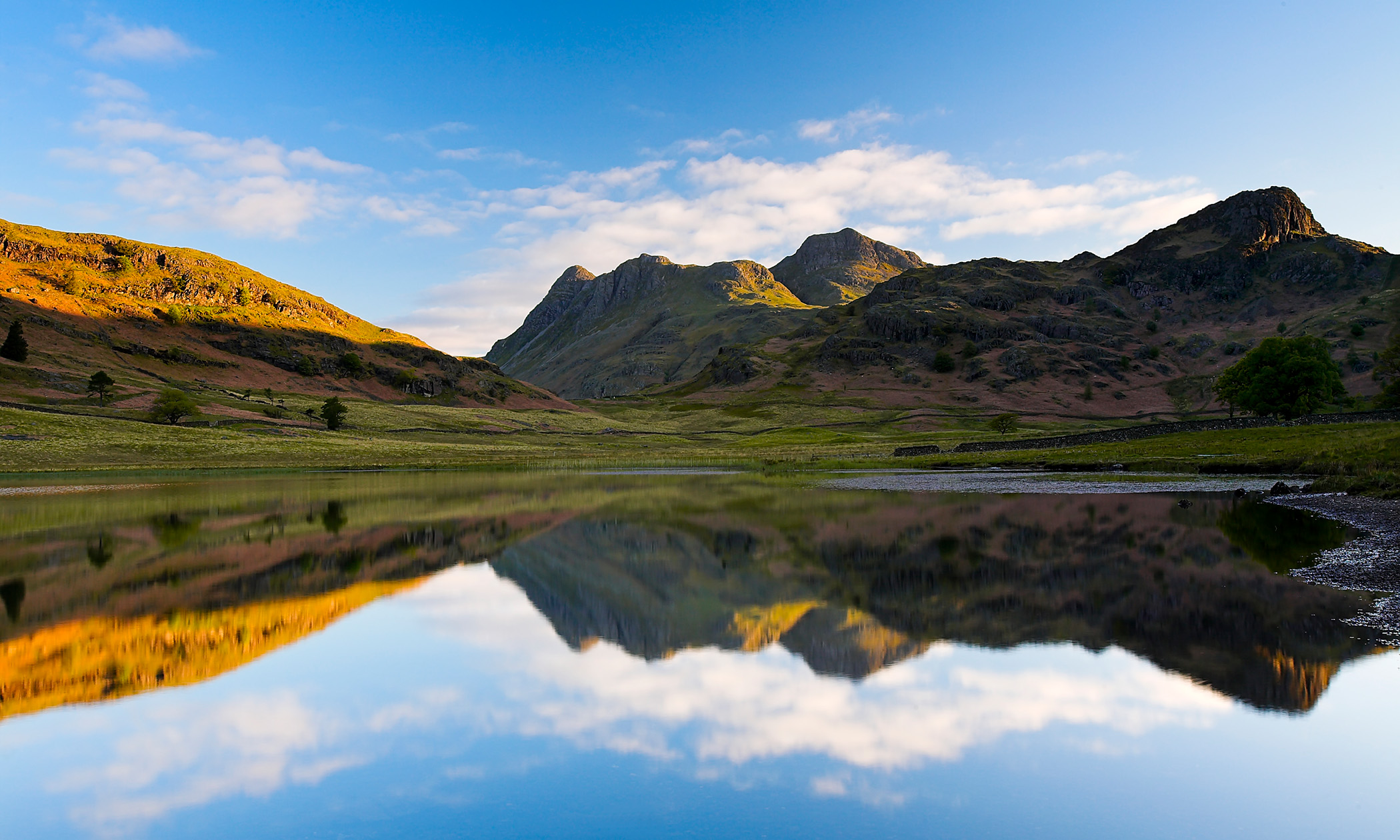 A scenic shot of a sunlit hillside beneath a blue sky, reflected in a lake in the foreground.