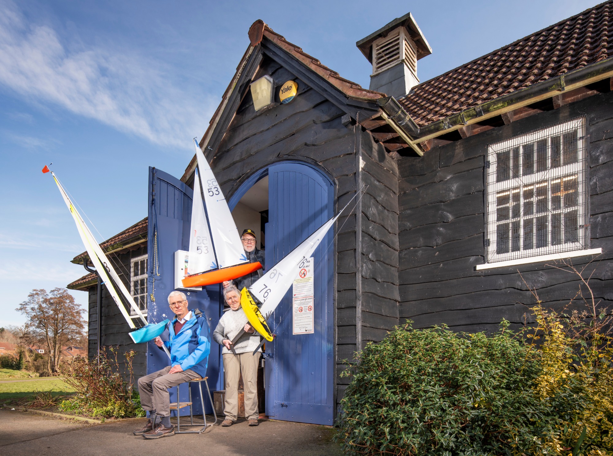 Three people holding model sailing boats outside a historic wooden boating house.