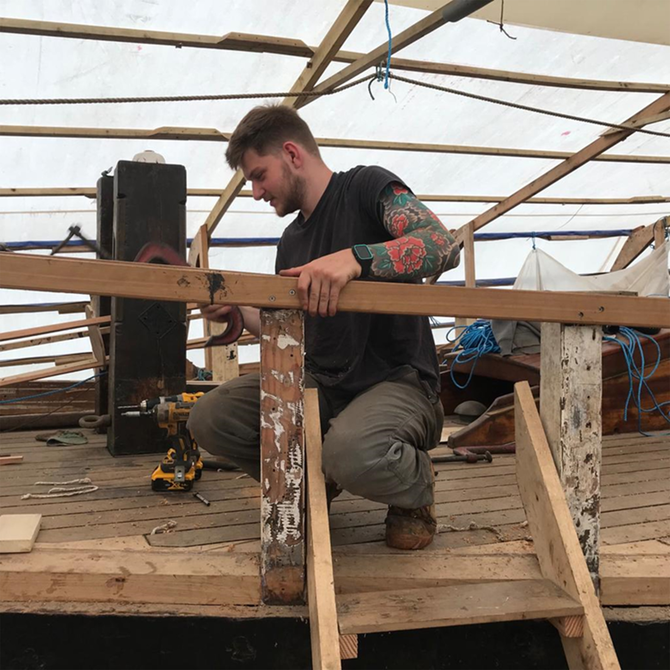 A volunteer works on a wooden structure. He is kneeling, using a tool to measure or adjust a wooden plank. The setting appears to be a workshop or construction site with scattered tools and materials.